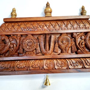 May include: A rectangular brown wooden wall hanging featuring intricate hand-carved details including two peacocks on the sides, a central U-shaped tilak symbol, a wheel, and a conch shell. Three small finials sit on top, and a small brass bell hangs below.