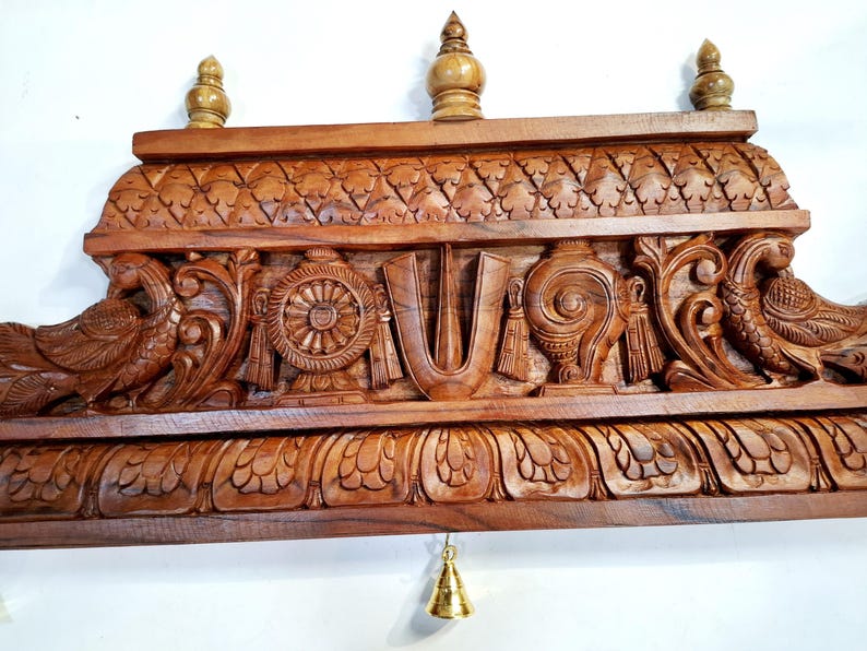 May include: A rectangular brown wooden wall hanging featuring intricate hand-carved details including two peacocks on the sides, a central U-shaped tilak symbol, a wheel, and a conch shell. Three small finials sit on top, and a small brass bell hangs below.