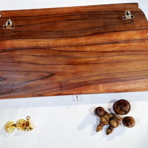 May include: A rectangular wooden wall shelf with a natural wood grain finish, featuring two metal keyhole hangers on the back and three small metal hooks along the bottom edge. Included are two brass-coloured bells and four wooden finial-style knobs.