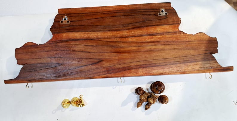 May include: A rectangular wooden wall shelf with a natural wood grain finish, featuring two metal keyhole hangers on the back and three small metal hooks along the bottom edge. Included are two brass-coloured bells and four wooden finial-style knobs.