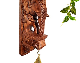 May include: A brown carved wooden wall mount featuring an elephant figure standing on a decorative bracket. The elephant has detailed textures and a beaded collar. A small, polished brass bell hangs from a hook at the bottom of the structure.