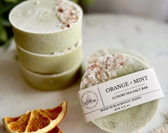 May include: A stack of four round, light green sea salt bars with pink salt crystals on top. One bar has a label that reads "ORANGE + MINT LUXURY SEA SALT BAR APX 4.6 oz." Two dried orange slices are in the foreground.