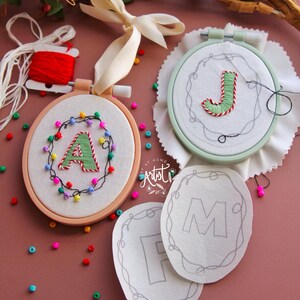 Peut inclure: Trois cerceaux à broder avec du tissu blanc et des contours noirs de lettres. Les lettres sont "A", "J" et "M". Un cerceau a un "A" vert brodé avec des perles colorées autour. Un autre cerceau a un "J" vert brodé. Le troisième cerceau a un "M" blanc avec un contour noir.