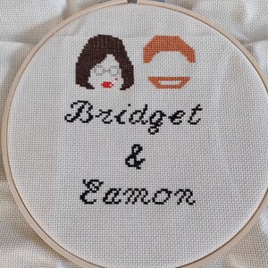 May include: A white embroidery hoop with a cross-stitch design featuring two cartoon faces and the text "Bridget & Eamon".