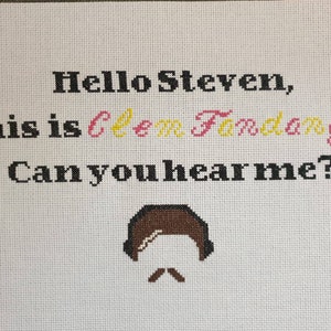 May include: Cross-stitch embroidery with the text "Hello Steven, this is Clem Fandango. Can you hear me?" in black, pink, and yellow thread. A brown cartoon figure with a mustache is stitched below the text.