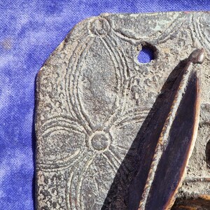 May include: Close-up of a weathered, bronze-colored decorative plaque with an embossed floral pattern. The plaque has a hole and a raised, pointed element. The background is a blue fabric.