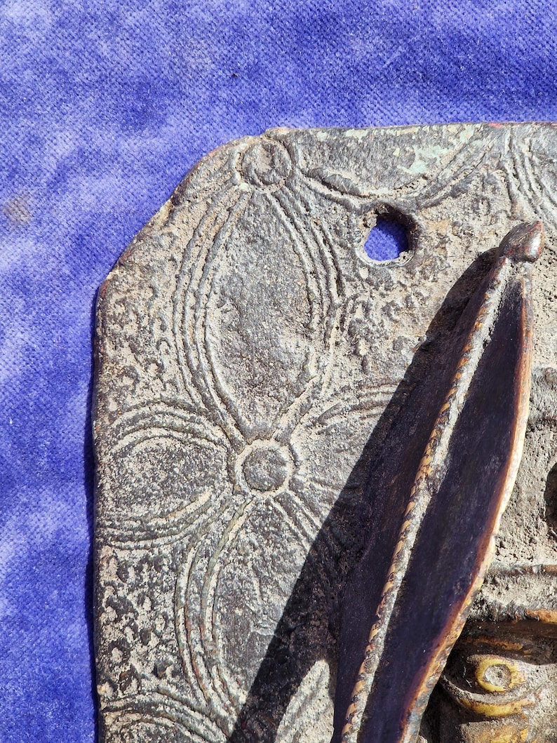 May include: Close-up of a weathered, bronze-colored decorative plaque with an embossed floral pattern. The plaque has a hole and a raised, pointed element. The background is a blue fabric.