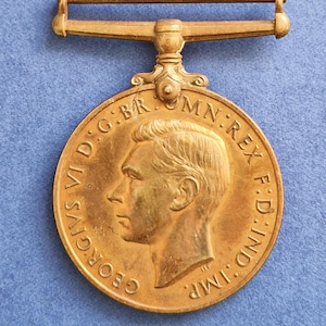 Puede incluir: Una medalla de color dorado con un perfil del rey Jorge VI en el anverso. La medalla tiene una cinta y la inscripción "GEORGIVS VID:GBR MN:REX F:D:IND:IMP." alrededor del borde.