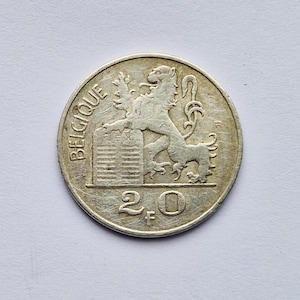 May include: A silver coin with the word "BELGIQUE" and a lion emblem. The coin also features the number "2.0" and the letters "F". The coin is circular and has a slightly worn appearance.