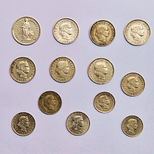 May include: A collection of 12 gold-colored Swiss coins with different dates and designs. The coins feature a woman's profile on one side and the text "CONFEDERATIO HELVETICA" on the other.