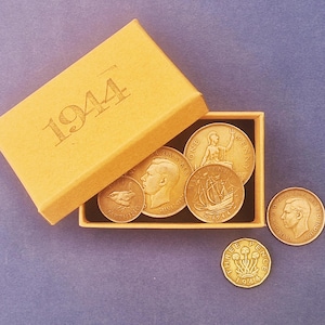Puede incluir: Una colección de monedas antiguas, incluyendo una moneda de tres peniques y una moneda de un penique, presentadas con una caja amarilla marcada "1944". Las monedas doradas presentan varios diseños y retratos.
