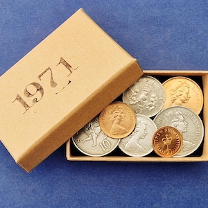 May include: A selection of vintage coins from 1971 presented in a small cardboard box. The box is tan with the year printed on the lid. The coins are silver and gold, featuring portraits and emblems.