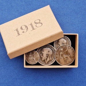 May include: A selection of antique coins presented in an open cardboard box. The box is beige and has the year "1918" printed on the lid. The coins are silver and bronze, displaying portraits and inscriptions.