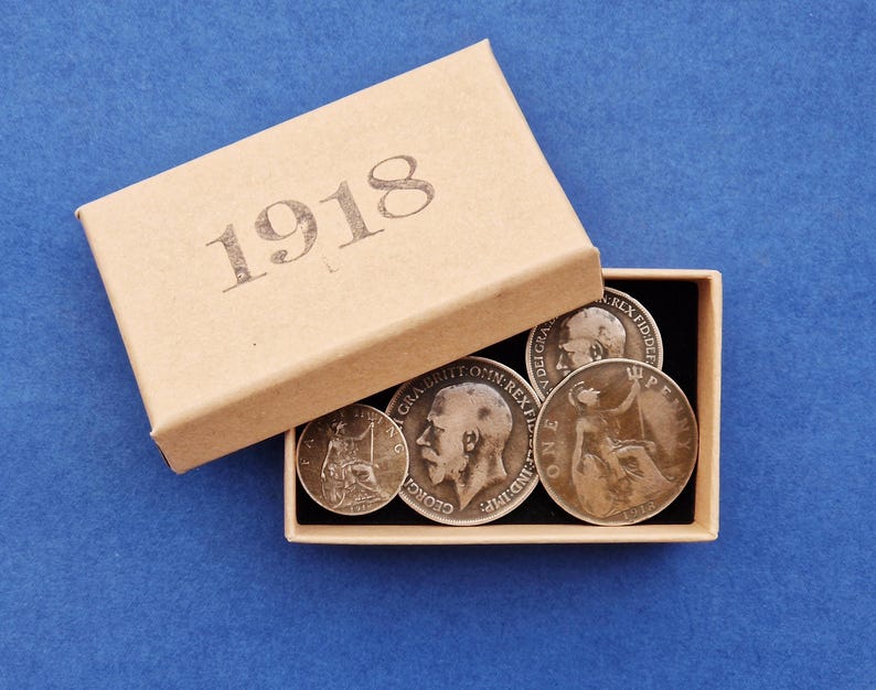 May include: A selection of antique coins presented in an open cardboard box. The box is beige and has the year "1918" printed on the lid. The coins are silver and bronze, displaying portraits and inscriptions.