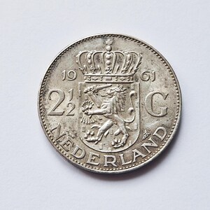 May include: A silver Dutch coin from 1961, featuring a crowned crest with a lion holding a sword and arrows. The coin has the text "NEDERLAND" around the edge and the denomination "2 1/2 G".