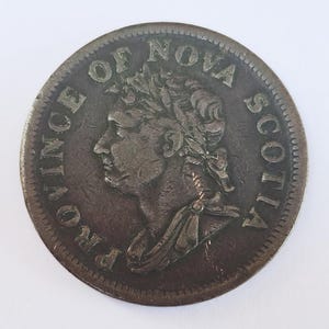 May include: A circular bronze-toned coin featuring a profile portrait of a monarch wearing a laurel wreath. The text PROVINCE OF NOVA SCOTIA is embossed along the upper edge of the coin, which shows signs of age and wear on its metallic surface.