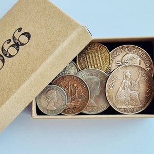 May include: A rectangular brown cardboard box with the text 1966 printed on the lid, containing a collection of vintage British coins. The coins feature various metallic tones, including copper and silver, with visible portraits and inscriptions.