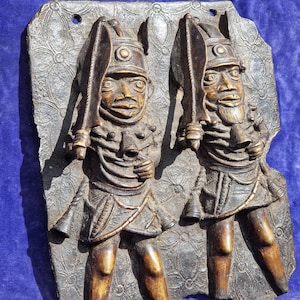 May include: A bronze relief sculpture featuring two figures holding swords. The figures wear helmets and ornate clothing, mounted on a textured background. The sculpture has a dark brown patina and is likely an antique or vintage piece.