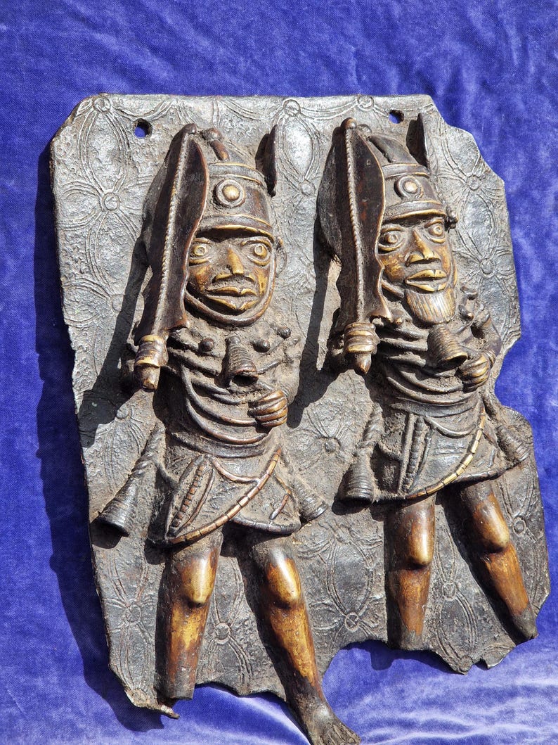 May include: A bronze relief sculpture featuring two figures holding swords. The figures wear helmets and ornate clothing, mounted on a textured background. The sculpture has a dark brown patina and is likely an antique or vintage piece.