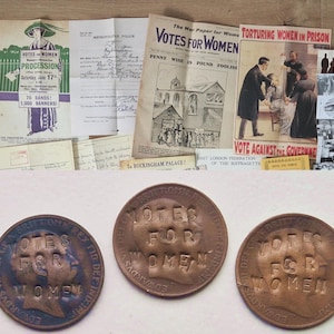 May include: A selection of historical items from the women's suffrage movement. The image shows coins, pamphlets, and posters, all bearing the slogan "Votes for Women." The items are aged, showing signs of use and historical significance.