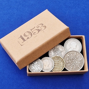 Puede incluir: Una colección de monedas antiguas de 1953 presentada en una pequeña caja de cartón abierta. La caja es de color beige con el año impreso en la tapa. Las monedas son plateadas y doradas, con diseños detallados.