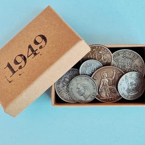 May include: A selection of old coins displayed in a small, open cardboard box. The box is a tan colour and has "1949" printed on the lid. The coins are silver and bronze, featuring different designs and portraits.