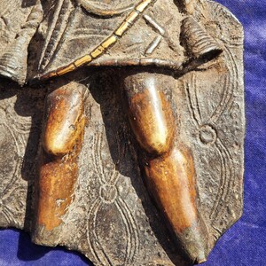 May include: A detailed, weathered bronze relief sculpture fragment depicting a figure with a spear and shield. The figure's legs and torso are visible, with intricate patterns and textures. The color palette includes shades of brown and gold.