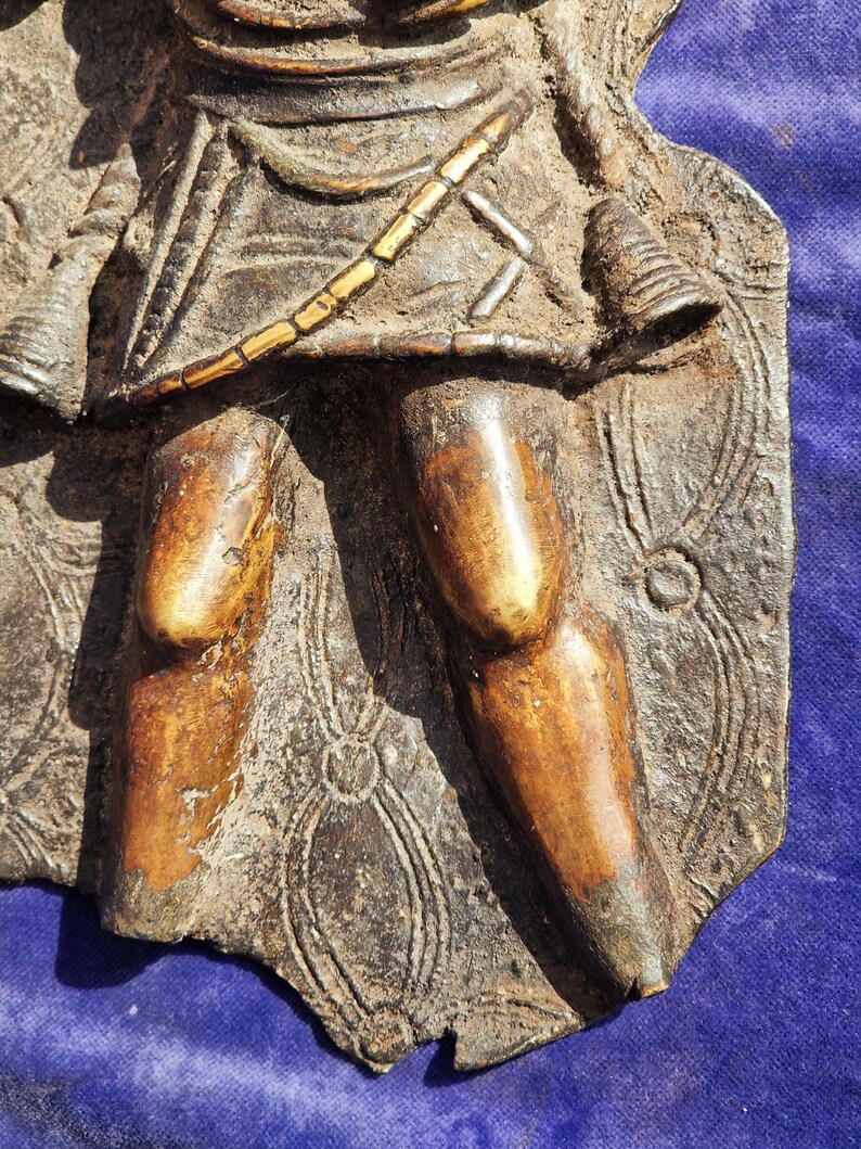 May include: A detailed, weathered bronze relief sculpture fragment depicting a figure with a spear and shield. The figure's legs and torso are visible, with intricate patterns and textures. The color palette includes shades of brown and gold.
