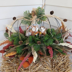 May include: A wire pumpkin frame decorated with faux greenery, pine cones, and two small fox figurines. The frame is sitting on a bed of straw.