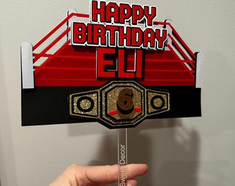 May include: A layered cake topper featuring a red and black wrestling ring design with the text HAPPY BIRTHDAY ELI in red and white. A gold glitter championship belt with the number 6 in the center is attached to the front on a clear stick.