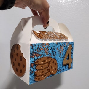 May include: A white cardboard cookie box with a blue and brown design. The box has a handle and says "Wesley" on the front. There are chocolate chip cookies on the front of the box and the number "4" on the side.