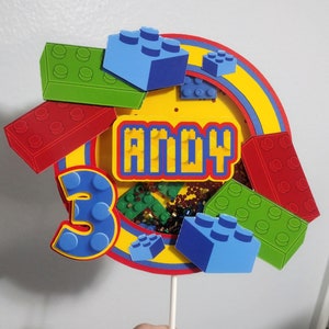May include: A colorful cake topper featuring a yellow circle with the name "ANDY" in pixelated letters. The number "3" is displayed in blue. Red, green, and blue toy brick accents surround the circle, with a rainbow arch.