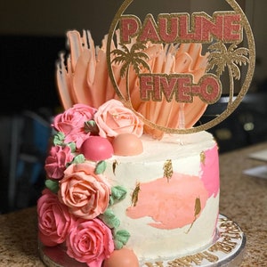 May include: A white cake decorated with pink roses, pink and gold accents, and a gold glitter circle topper that says "PAULINE FIVE-O".