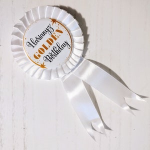 May include: A white rosette badge with a pleated edge and two flowing ribbons. The center circle features the text "Ilarinyz GOLDEN Birthday" in gold and black, with star accents. A celebratory accessory.