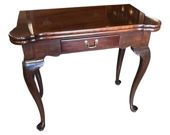 Puede incluir: Una mesa auxiliar de madera marrón oscuro con un cajón y patas curvas. La mesa tiene una parte superior rectangular con bordes redondeados y un tirador de cajón de latón. Las patas están elegantemente curvadas, lo que se suma a la estética antigua de la mesa. La mesa mide aproximadamente 76 cm de alto.