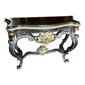 May include: An antique console table with a black top and ornate gold and black detailing. The table features a curved design with decorative carvings and a gold accent along the edges. The legs are curved and the table has a classic, vintage aesthetic.