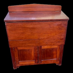 May include: A vintage wooden cabinet with a hinged top and two doors. The cabinet is made of dark wood with a reddish hue and has a simple design.