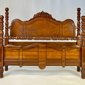 May include: A medium-brown wooden bed frame featuring a tall headboard with a carved crest, a matching footboard, and four turned posts with spherical finials. The frame includes a horizontal spindle rail connecting the posts at the headboard.
