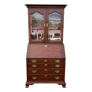 May include: A dark wood secretary desk with a hutch. The hutch has two glass doors with brass hardware. Below the hutch is a slanted desk surface and four drawers with brass hardware. The desk has a traditional design.