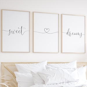 May include: Three framed prints with the words "sweet", a heart, and "dreams" in black calligraphy on a white background. The prints are hanging on a white wall above a bed with white bedding and a cream-colored throw blanket.