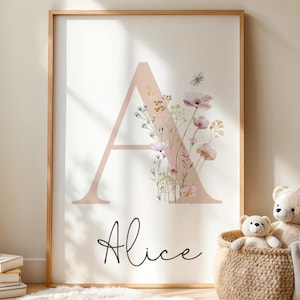 Può includere: Stampa incorniciata con una grande lettera "A" color rosa pallido adornata da fiori ad acquerello in tenui tonalità rosa e gialle. Il nome "Alice" è scritto in un'elegante grafia sottostante. L'opera è su sfondo bianco.