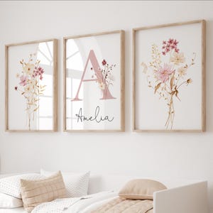 Könnte beinhalten: Drei gerahmte Kunstdrucke mit Blumenmotiven. Die Drucke zeigen Aquarellbouquets in Rosa-, Creme- und Goldtönen. Der mittlere Druck hat einen großen rosa Buchstaben "A" mit dem Namen "Amelia" in Schreibschrift. Die Rahmen sind aus hellem Holz.