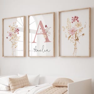 Peut inclure: Trois impressions d'art floral encadrées. Les impressions présentent des bouquets à l'aquarelle dans des tons roses, crème et dorés. L'impression centrale comporte un grand "A" rose avec des accents floraux et le nom "Amelia" en écriture cursive. Les cadres sont en bois clair.