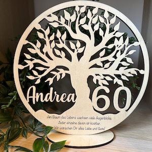 Könnte beinhalten: Ein hölzernes, kreisförmiges, lasergeschnittenes Lebensbaum-Design mit dem Namen "Andrea" und der Zahl "60". Der Baum ist vor einem dunklen Hintergrund platziert, mit Text unten. Der Sockel ist ebenfalls aus Holz.
