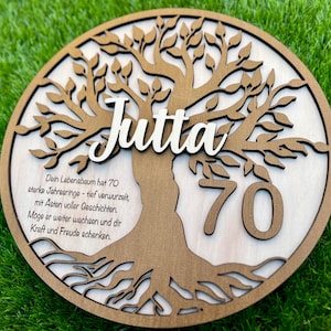 Könnte beinhalten: Runde Holzplakette mit Baumdesign, dem Namen "Jutta" in weißer Schrift und der Zahl "70". Deutscher Text ist enthalten. Die Plakette besteht aus hellem und dunklem braunem Holz mit natürlichem Finish.