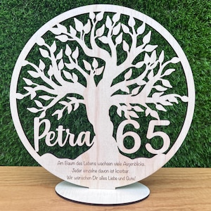 May include: A wooden tree of life decoration with the name "Petra" and the number "65" cut out. The tree is inside a circle, and the base has text in German. The decoration is on a wooden stand.
