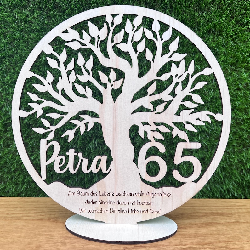 May include: A wooden tree of life decoration with the name "Petra" and the number "65" cut out. The tree is inside a circle, and the base has text in German. The decoration is on a wooden stand.