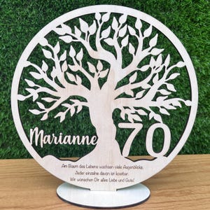 May include: A wooden, laser-cut tree of life design with the name "Marianne" and the number "70". The tree is inside a circular frame. The base has text in German. The piece is displayed on a wooden stand.