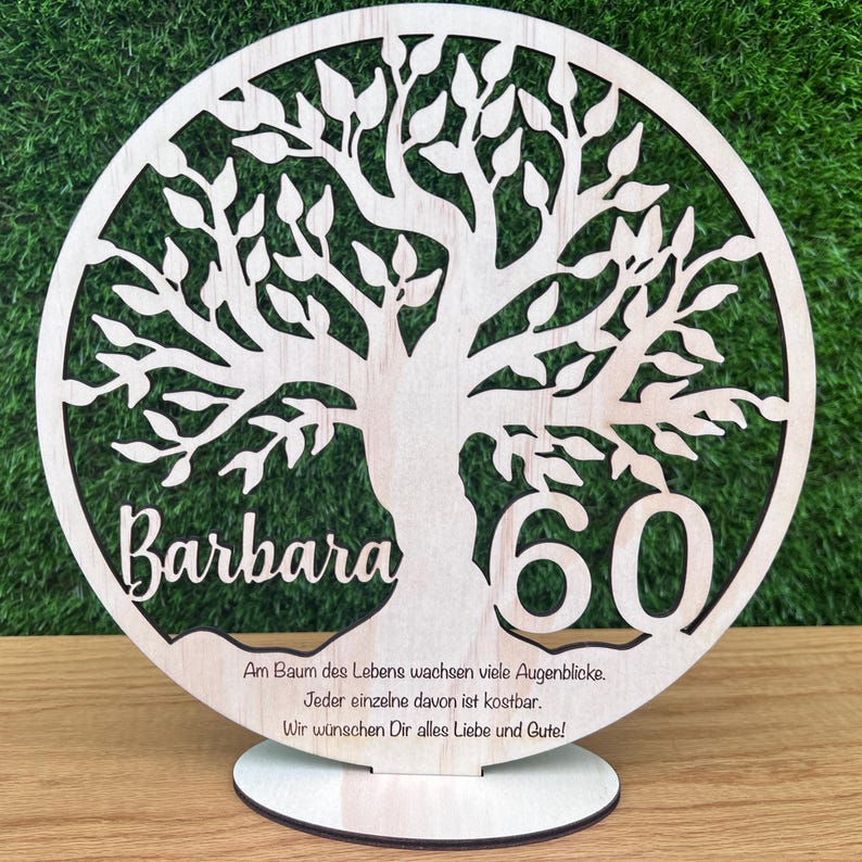 May include: A wooden, laser-cut tree of life decoration. The design includes the name "Barbara" and the number "60". The base has text in German. The tree is set against a green background.