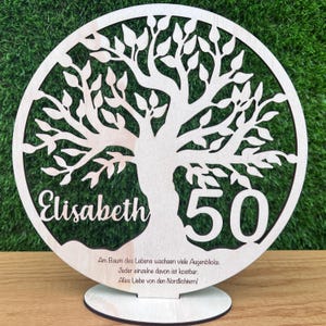 May include: A wooden, circular tree of life decoration with the name "Elisabeth" and the number "50" cut out. The tree has many leaves and is mounted on a round base. Text at the bottom reads in German.